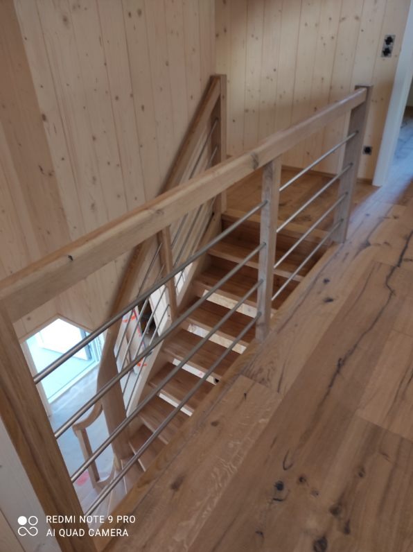 Jasne, drewniane schody z metalową balustradą, widziane z góry. Naturalne oświetlenie podkreśla strukturę drewna na stopniach i podłodze. Minimalistyczny design.