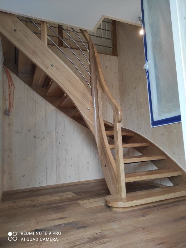 Jasne, drewniane schody z metalową balustradą, zakręcające w prawo, na tle boazerii i drewnianej podłogi. Widoczne detale wykończenia i naturalne usłojenie drewna.