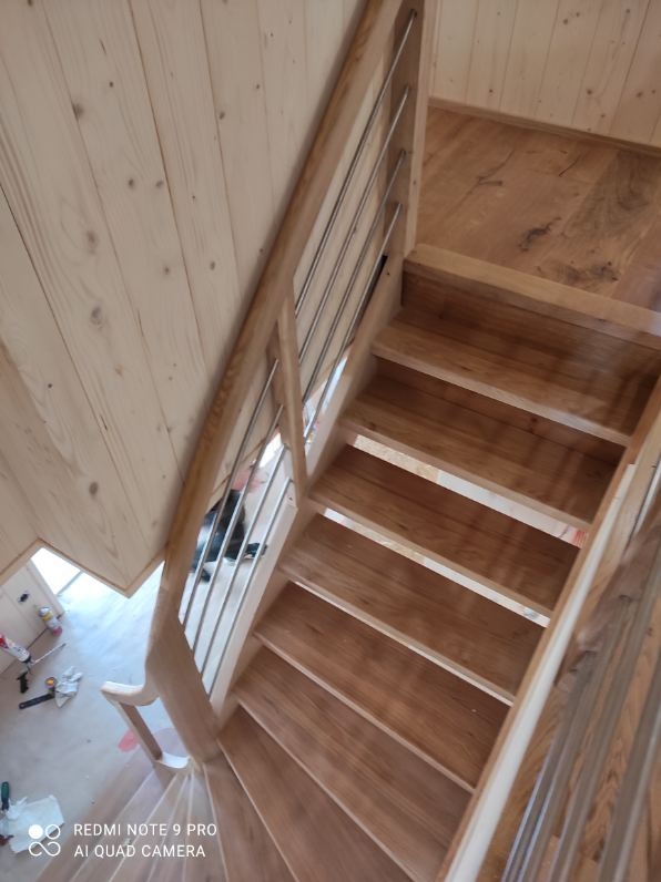 Jasne, drewniane schody z metalową balustradą, widok z góry. Naturalne oświetlenie podkreśla strukturę drewna. Ściany wykończone boazerią.