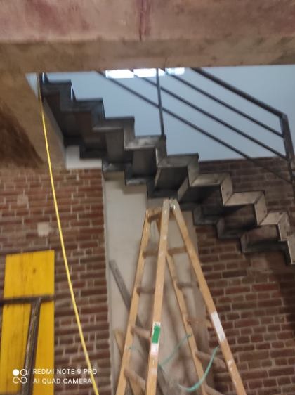 Surowe schody metalowe w trakcie montażu, widoczne z bliska, z drewnianą drabiną i fragmentem ceglanej ściany w tle. Elementy konstrukcyjne w stanie nieukończonym.