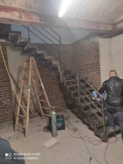 Montaż metalowych schodów z balustradą w surowym wnętrzu z ceglaną ścianą. Widoczna drabina i butla spawalnicza, sugerujące trwające prace wykończeniowe.