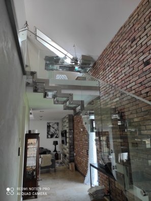 Szklane schody z balustradą w nowoczesnym wnętrzu z ceglaną ścianą. Widok z dołu, podkreślający minimalistyczny design i transparentność konstrukcji.