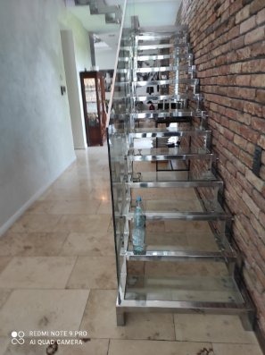 Nowoczesne schody ze stali nierdzewnej z szklaną balustradą, na tle ceglanej ściany. Widoczna butelka wody na jednym ze stopni. Jasna podłoga z płytek.