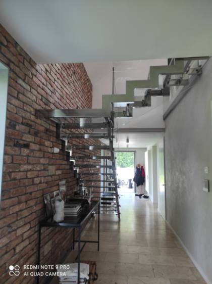 Nowoczesne schody metalowe z minimalistyczną konstrukcją, przylegające do ceglanej ściany w jasnym wnętrzu. Widoczne elementy dekoracyjne i oświetlenie.