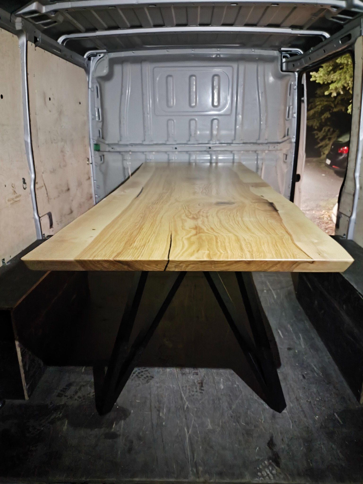 Blat stołu z jasnego drewna z widocznym usłojeniem i czarnymi nogami, umieszczony w przestrzeni ładunkowej samochodu dostawczego. Nietypowe ujęcie.