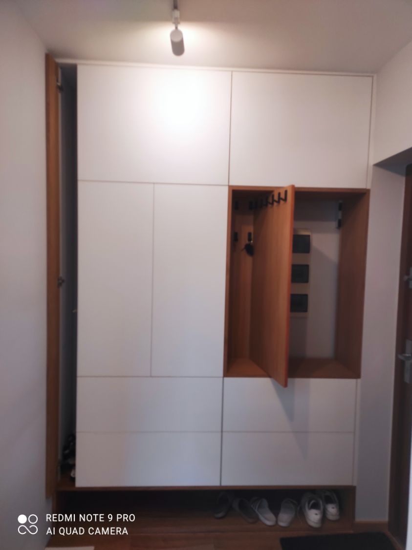 Biała szafa wnękowa z drewnianymi akcentami i otwartą półką na buty. Otwarta szafka z wieszakami i panelem elektrycznym w tle. Nowoczesny design.