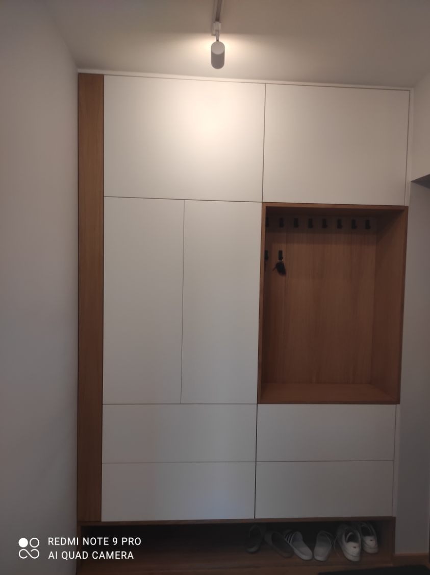 Biała szafa wnękowa z drewnianymi akcentami i otwartą przestrzenią na wieszaki oraz buty, oświetlona punktowo, minimalistyczny design.