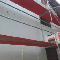 Ocieplanie elewacji budynku styropianem, widoczne rusztowanie z czerwonymi podestami i paczki styropianu z oznaczeniami producenta na rusztowaniu.