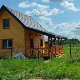 Waldemar Stasik - Żółty dom szkieletowy z ciemnym dachem i drewnianą werandą, otoczony zielenią i ogrodzeniem. Błękitne niebo z obłokami w tle.
