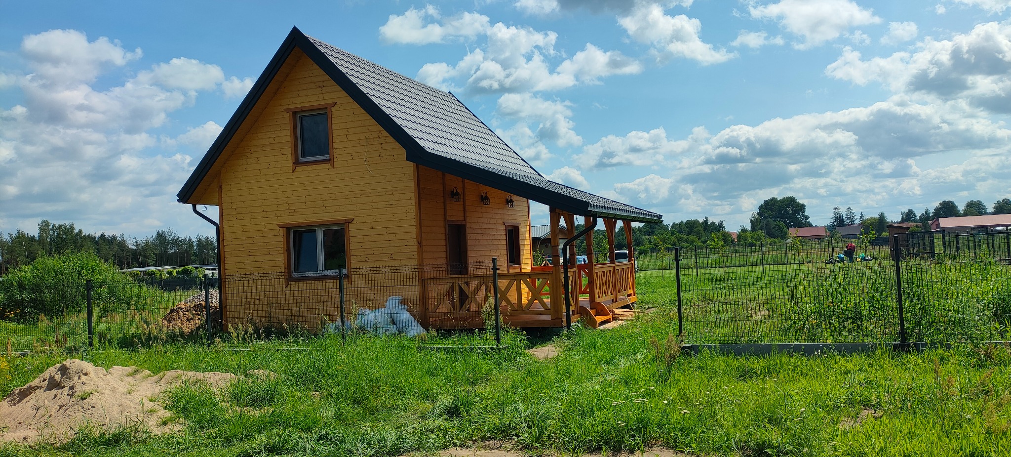 Żółty dom szkieletowy z ciemnym dachem i drewnianą werandą, otoczony zielenią i ogrodzeniem. Błękitne niebo z obłokami w tle.