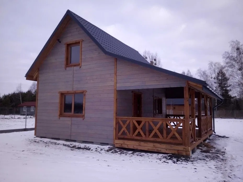 Zimowy krajobraz z szarym domem szkieletowym z drewnianą werandą i ciemnym dachem. Widok z boku na tle zaśnieżonego terenu i drzew.