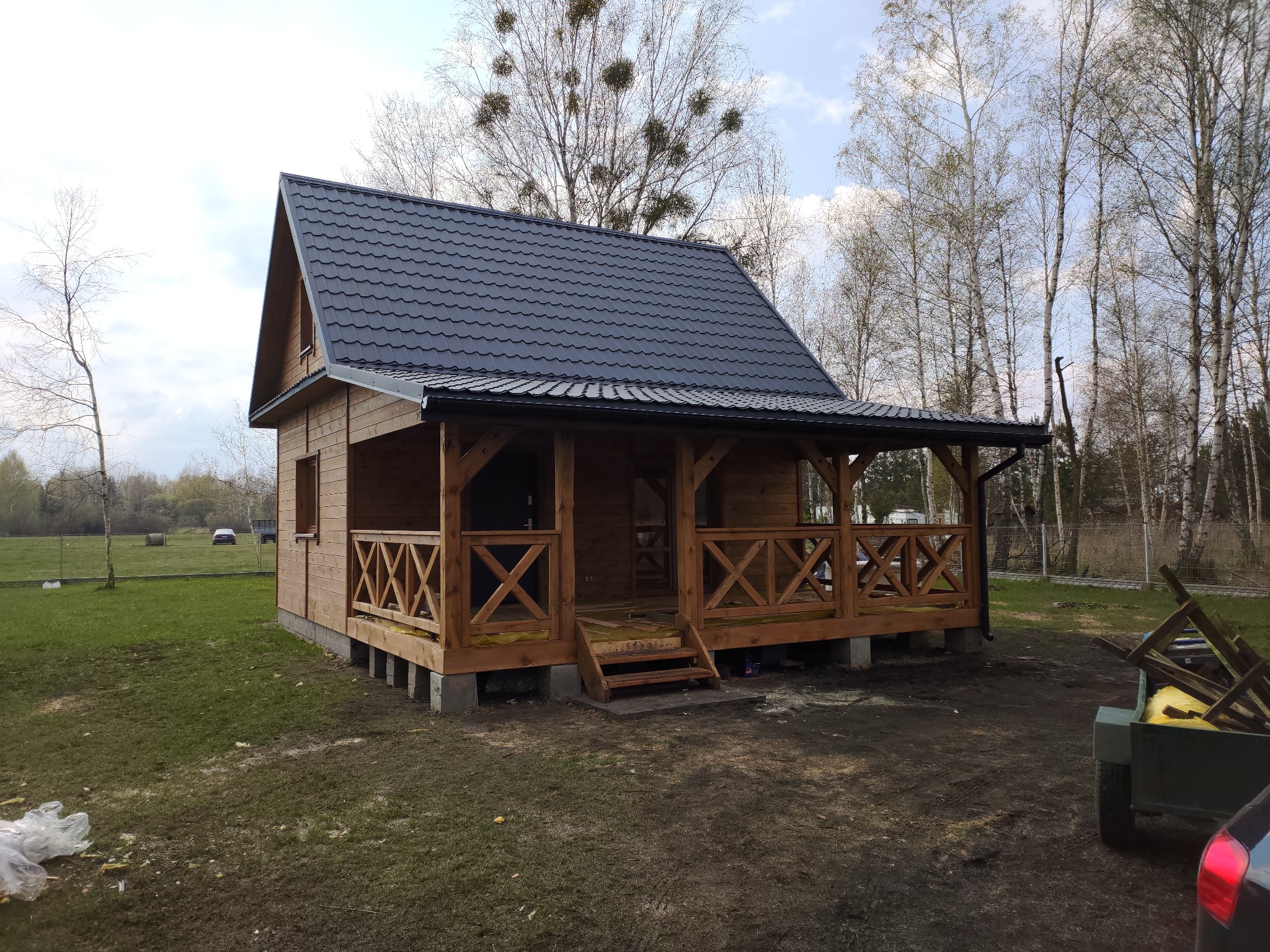 Niewielki, parterowy dom szkieletowy z zadaszonym tarasem, wykończony drewnem, z ciemnym dachem z blachodachówki, osadzony na betonowych bloczkach.