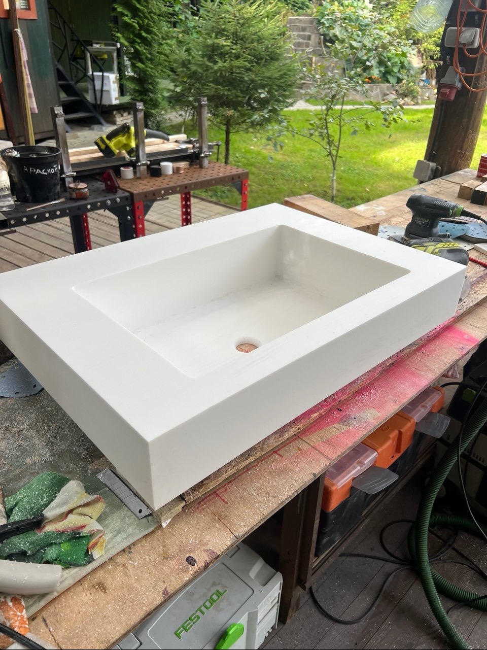 Biała, prostokątna umywalka z konglomeratu na warsztatowym stole, otoczona narzędziami i akcesoriami. W tle zieleń ogrodu i konstrukcja metalowa.