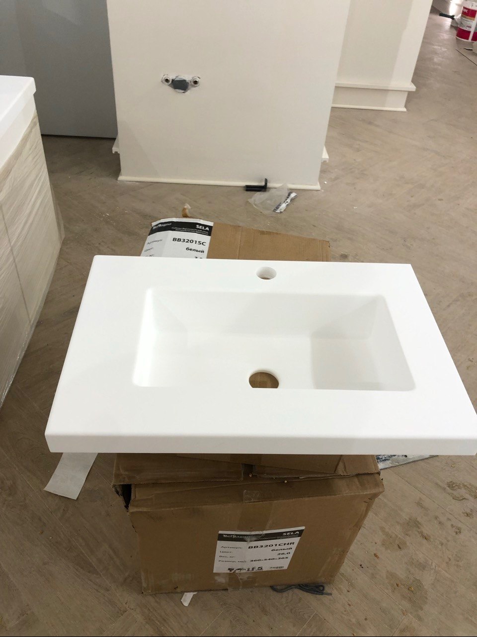 Biała umywalka nablatowa SELA BB3201SC na kartonie, przygotowana do montażu. Widoczne przyłącza wodne w ścianie. Minimalistyczny design, surowe wykończenie.