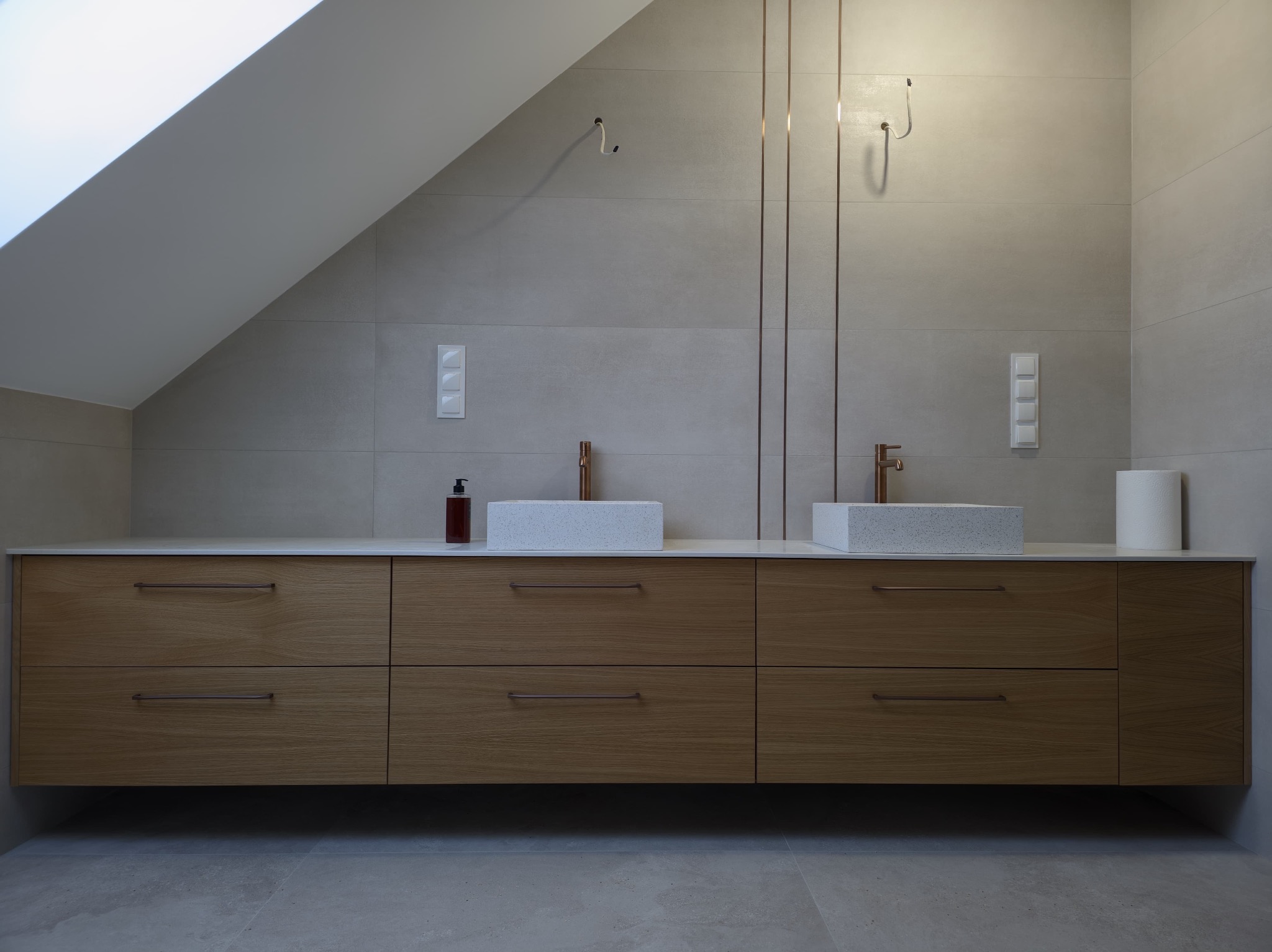 Nowoczesna łazienka na poddaszu z drewnianą szafką pod umywalki, kamiennymi blatami i miedzianą armaturą. Minimalistyczny design, szare płytki i subtelne oświetlenie.