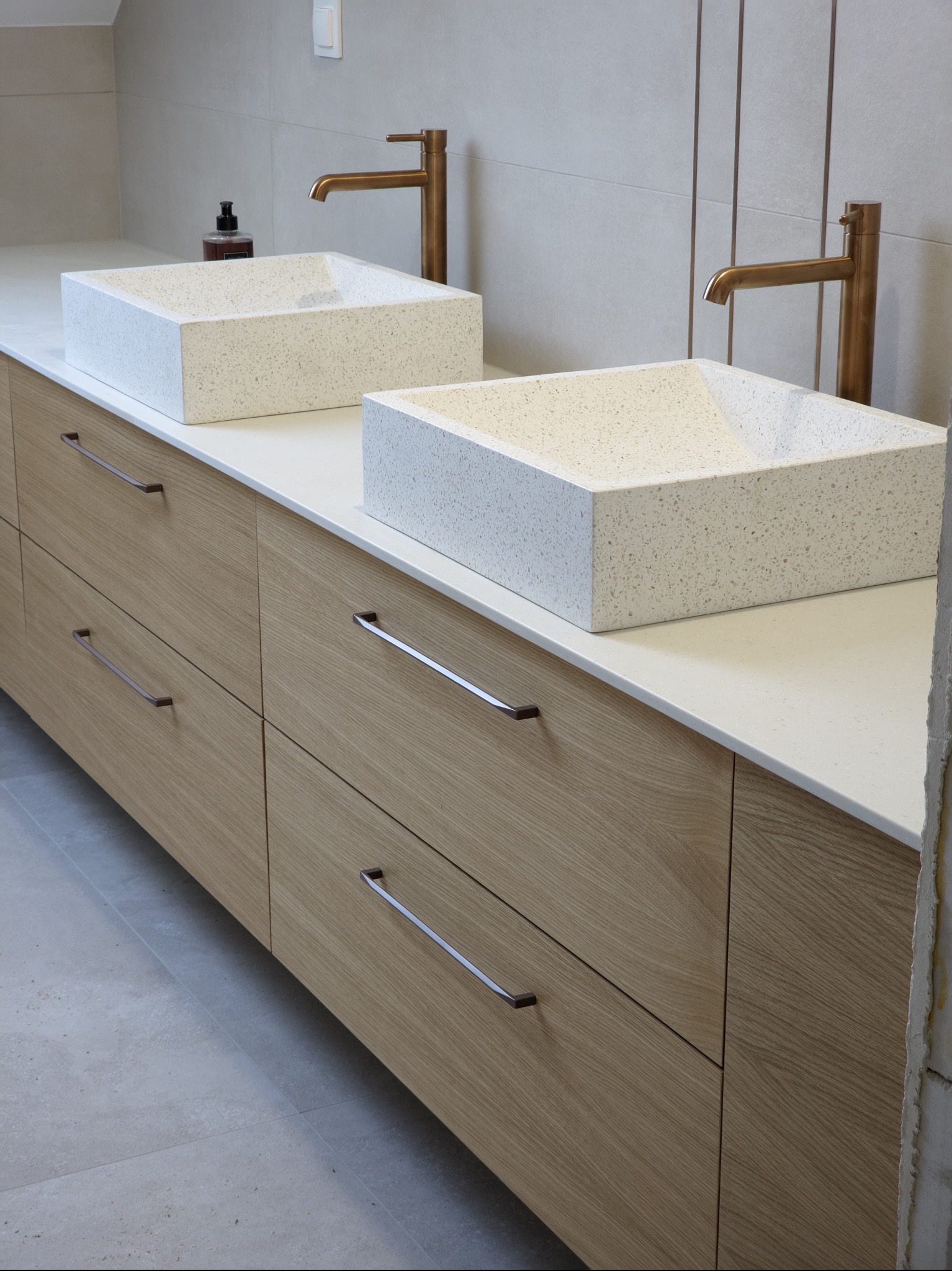 Nowoczesna łazienka z dwiema kwadratowymi umywalkami z konglomeratu na drewnianej szafce z podłużnymi uchwytami i miedzianą armaturą na tle jasnych płytek.