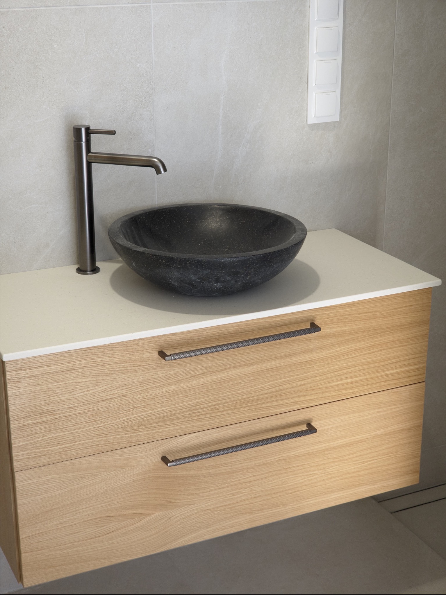 Łazienkowa szafka z jasnego drewna z dwoma szufladami i blatem z konglomeratu, na którym stoi czarna, okrągła umywalka i nowoczesna bateria w kolorze grafitowym.