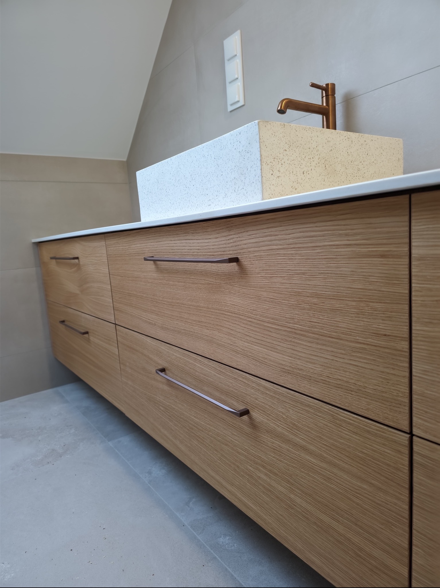 Nowoczesna szafka łazienkowa z drewnianymi frontami, umywalką z konglomeratu i miedzianą armaturą. Minimalistyczny design, jasne płytki w tle.