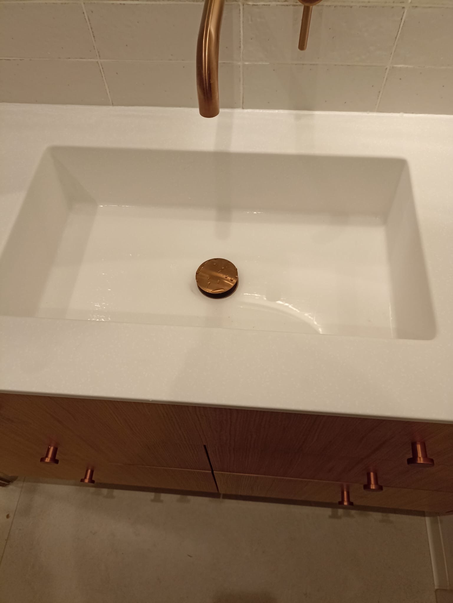 Biała umywalka z blatem z drewnopodobnego materiału i miedzianą armaturą. Widoczny korek spustowy z detalami. Minimalistyczny design.