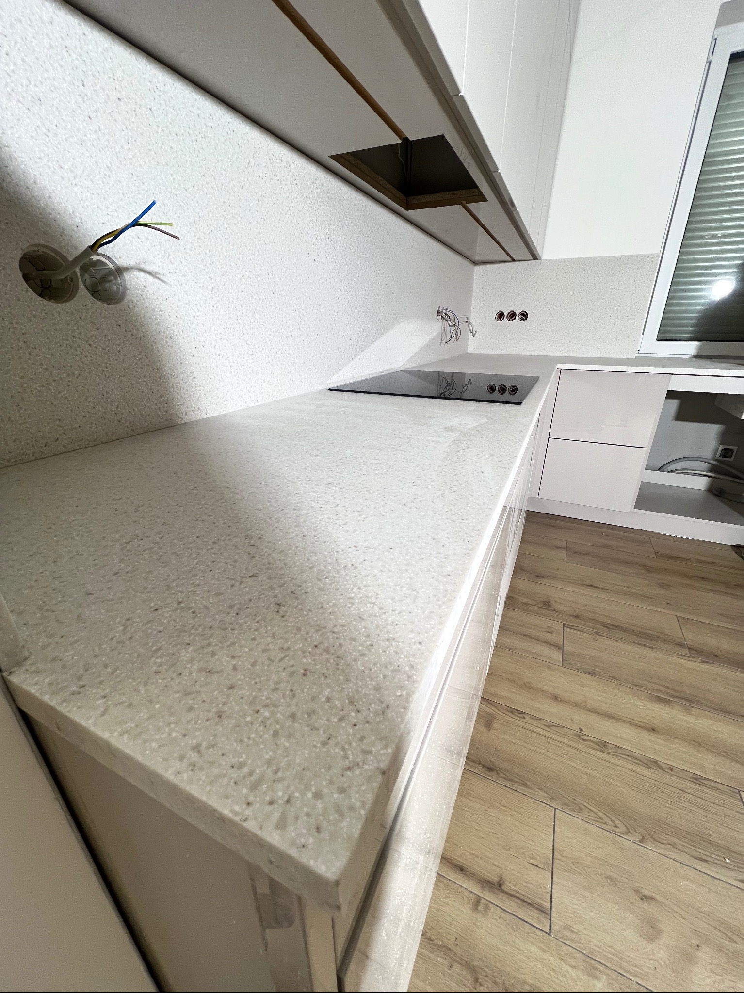 Jasny blat z konglomeratu w kuchni z niedokończonymi instalacjami elektrycznymi. Widoczne gniazdka i przewody. Ujęcie z perspektywy podłogi, ukazujące strukturę blatu.
