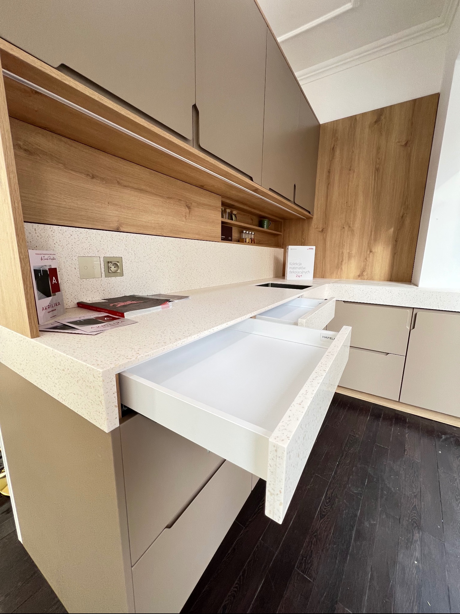 Jasna kuchnia z blatem z konglomeratu i otwartą szufladą. Widoczne drewniane wykończenia i ciemna podłoga. Na blacie materiały reklamowe Akrilika.
