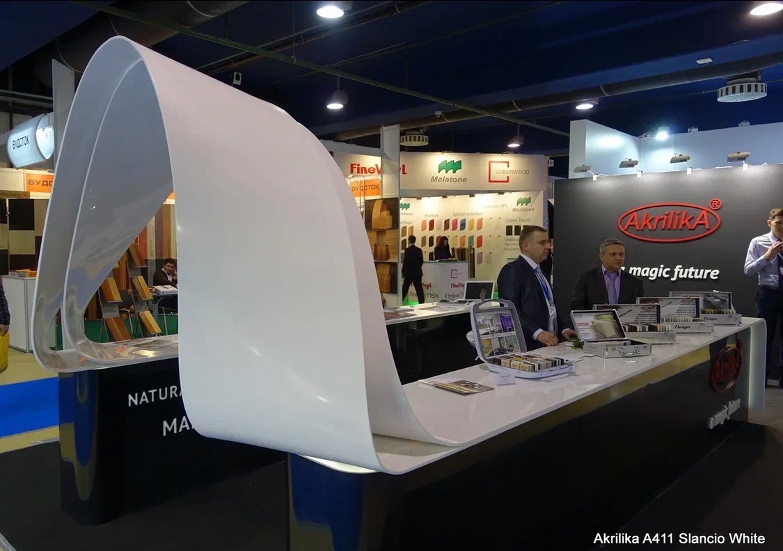 Ekspozycja stoiska targowego z próbkami materiałów wykończeniowych, w tym blatów i frontów meblowych. Widoczny element z białego akrylu w formie łuku.