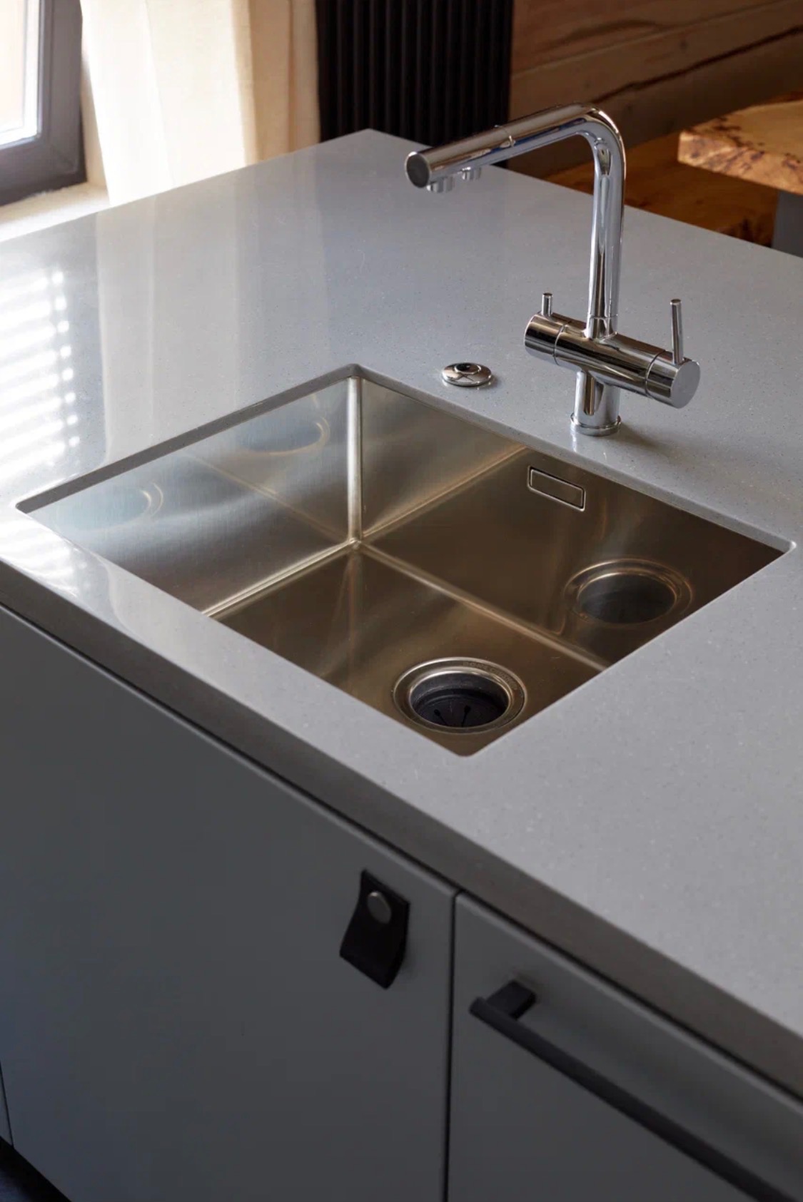 Szary blat z konglomeratu z nowoczesnym, chromowanym kranem i wpuszczanym, dwukomorowym zlewem ze stali nierdzewnej. Minimalistyczne, ciemne uchwyty szafek.
