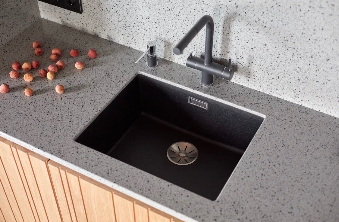 Szary blat z konglomeratu z czarnym zlewem i baterią. Na blacie leżą owoce liczi. Ujęcie z góry, prezentujące wykończenie kuchni.