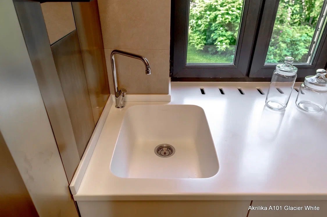 Biały, minimalistyczny zlew wkomponowany w blat kuchenny z akryliku A101 Glacier White, z widoczną baterią i oknem z zielonym widokiem na zewnątrz.