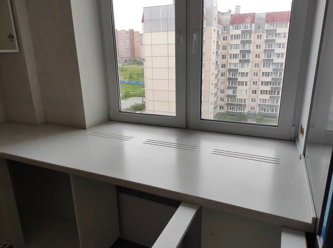 Jasny parapet z konglomeratu z trzema rzędami otworów wentylacyjnych, zamontowany pod oknem z widokiem na budynki mieszkalne. Widoczny grzejnik i konstrukcja pod parapetem.