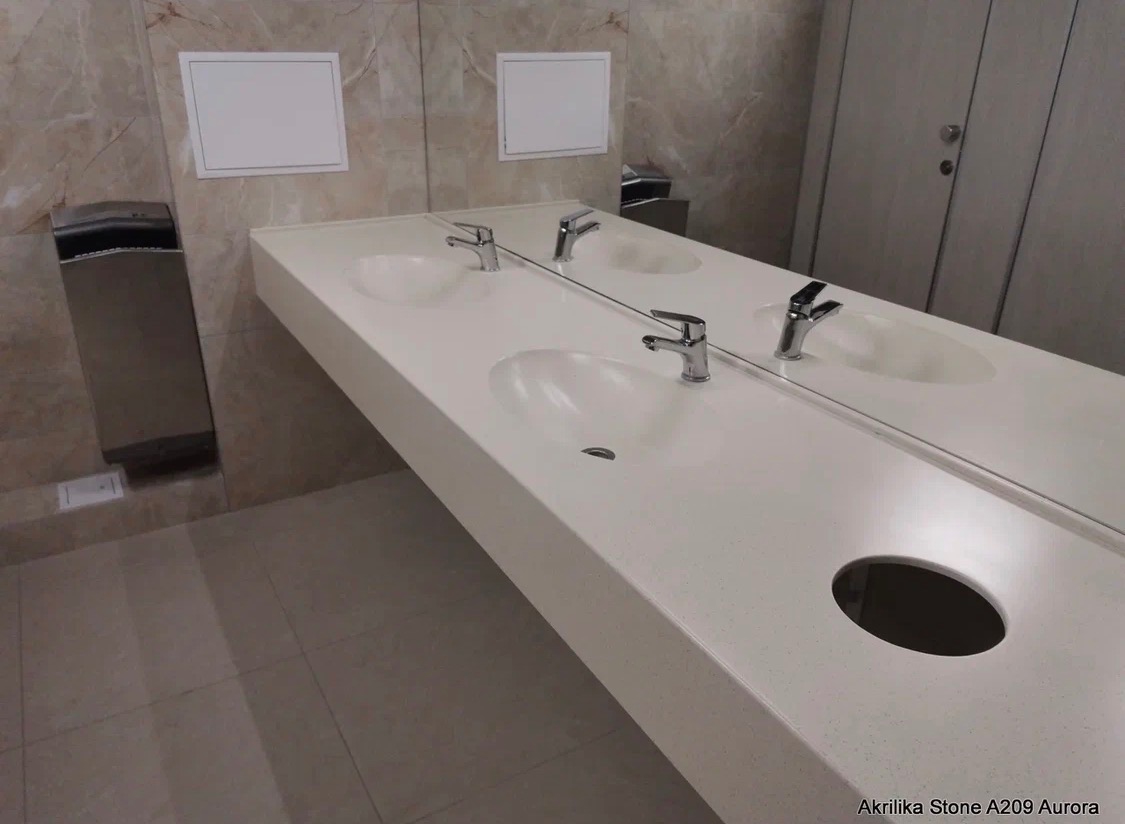 Długi, biały blat z konglomeratu z dwiema umywalkami i bateriami, zamontowany w publicznej toalecie. Widoczne lustro, dozownik i fragment ściany z płytkami.