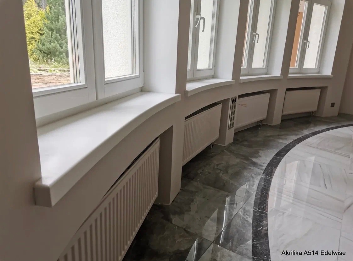 Białe, zaokrąglone parapety z konglomeratu pod oknami, z grzejnikami wnękowymi. Szara, marmurkowa podłoga z ciemnym obramowaniem. Widok na zieleń za oknem.