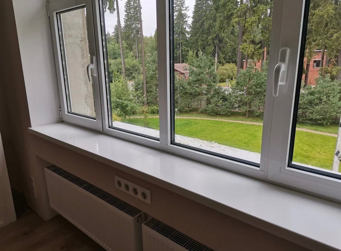 Biały parapet z konglomeratu pod oknem z widokiem na zieleń. Nowoczesny design, gładka powierzchnia, widoczne gniazdka elektryczne i grzejnik pod parapetem.