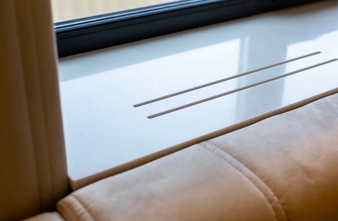 Biały parapet z konglomeratu z dwoma szczelinami wentylacyjnymi, zamontowany przy oknie, przylegający do beżowej sofy. Minimalistyczny design, jasne wnętrze.