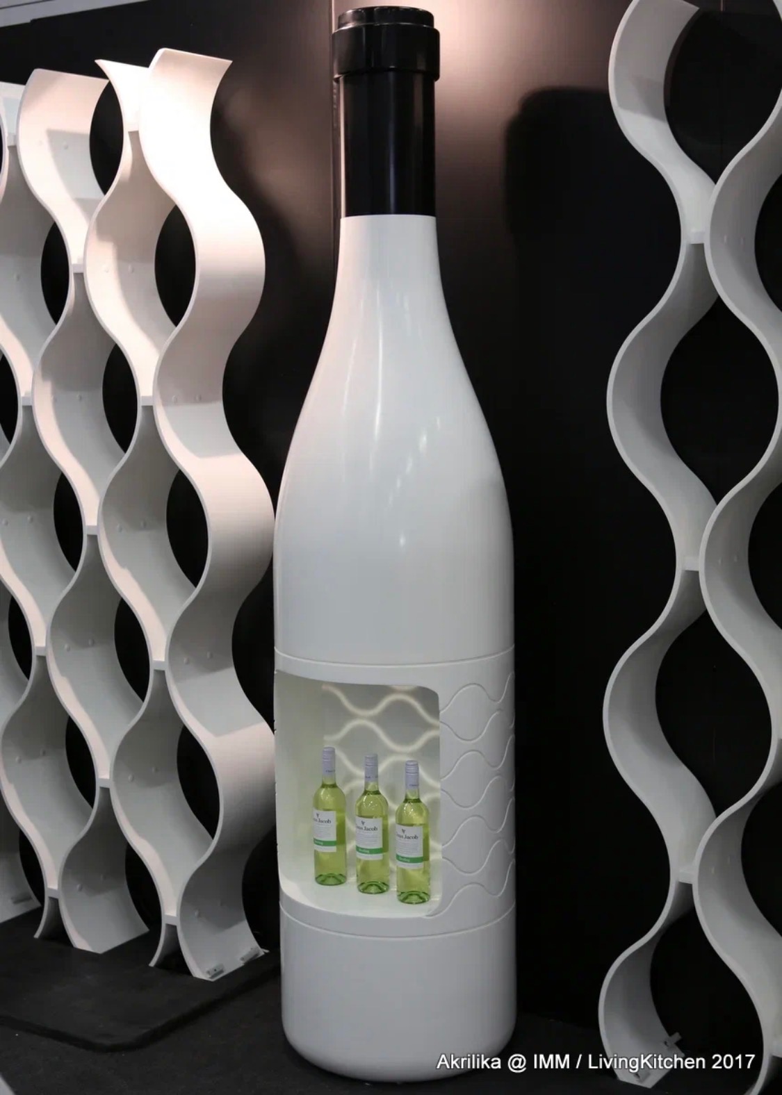 Biały regał na wino w kształcie butelki, z trzema butelkami w środku, obok ażurowe półki na wino o falistym kształcie na czarnym tle. Nowoczesny design.
