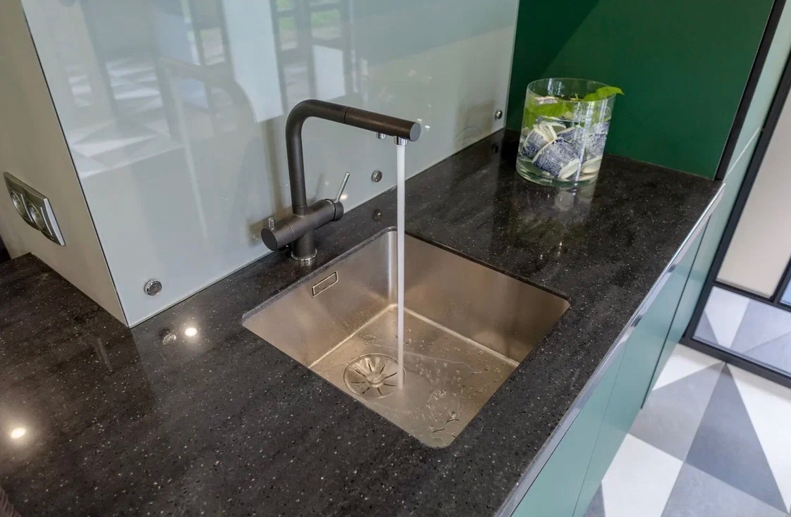 Czarny, błyszczący blat z konglomeratu z metalowym zlewem i nowoczesną baterią, z której leci woda. W tle szklany panel i dekoracja z roślin w szklanym wazonie.