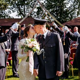 Piotr Jędrzejewski Fotograf Ślubny i Rodzinny - Pocałunek pary młodej w mundurach pod szpalerem szabel. Panna młoda w koronkowej sukni trzyma bukiet. Uroczystość na zewnątrz, w tle drzewa i czerwone krzesła.
