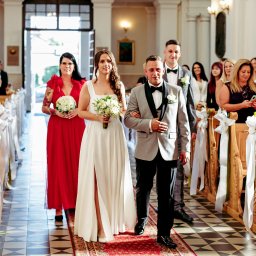 Piotr Jędrzejewski Fotograf Ślubny i Rodzinny - Para młoda prowadzona do ołtarza w kościele, goście obserwują i robią zdjęcia. Dekoracje z białych wstążek na ławkach. Panna młoda w sukni z rozcięciem.