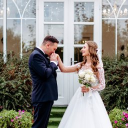 Piotr Jędrzejewski Fotograf Ślubny i Rodzinny - Elegancka para młoda: pan młody całuje dłoń uśmiechniętej panny młodej z bukietem białych róż na tle białych drzwi i zieleni.