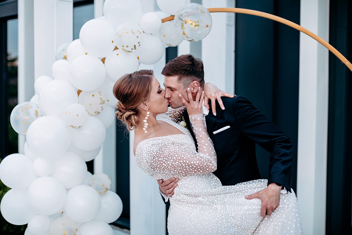 Pocałunek pary młodej na tle białych balonów i złotej konstrukcji. Panna młoda w sukni z długim rękawem w kropki, pan młody w czarnym garniturze. Ujęcie z bliska.