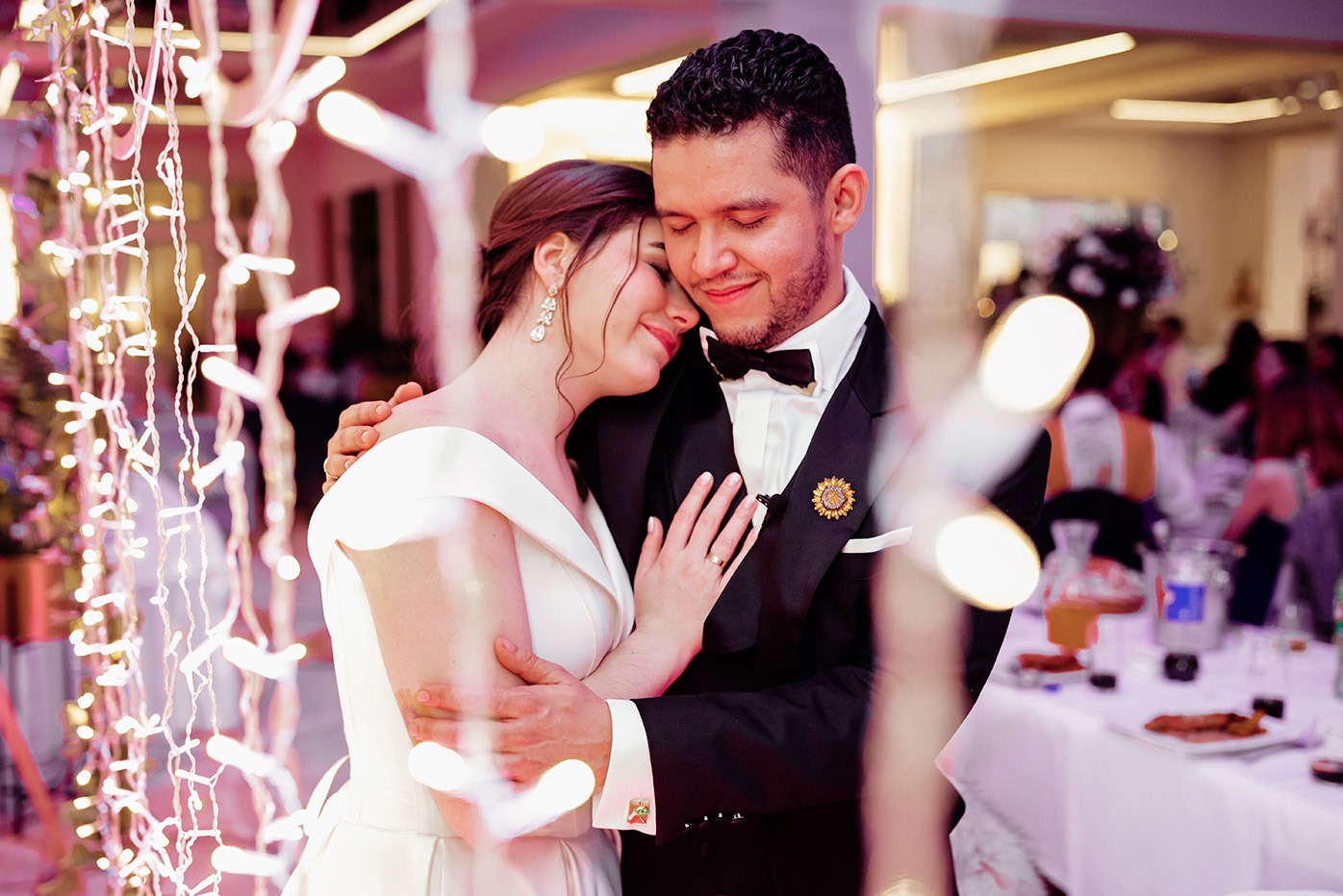 Uśmiechnięta para młoda w objęciach, panna młoda w białej sukni, pan młody w czarnym smokingu z ozdobną broszką, ciepłe oświetlenie i dekoracje w tle.