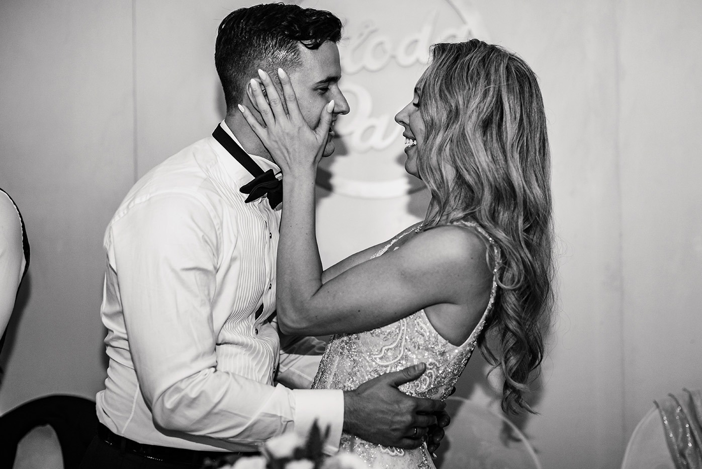 Czarno-białe zdjęcie uśmiechniętej pary młodej w bliskim ujęciu. Panna młoda dotyka twarzy pana młodego. W tle dekoracja.