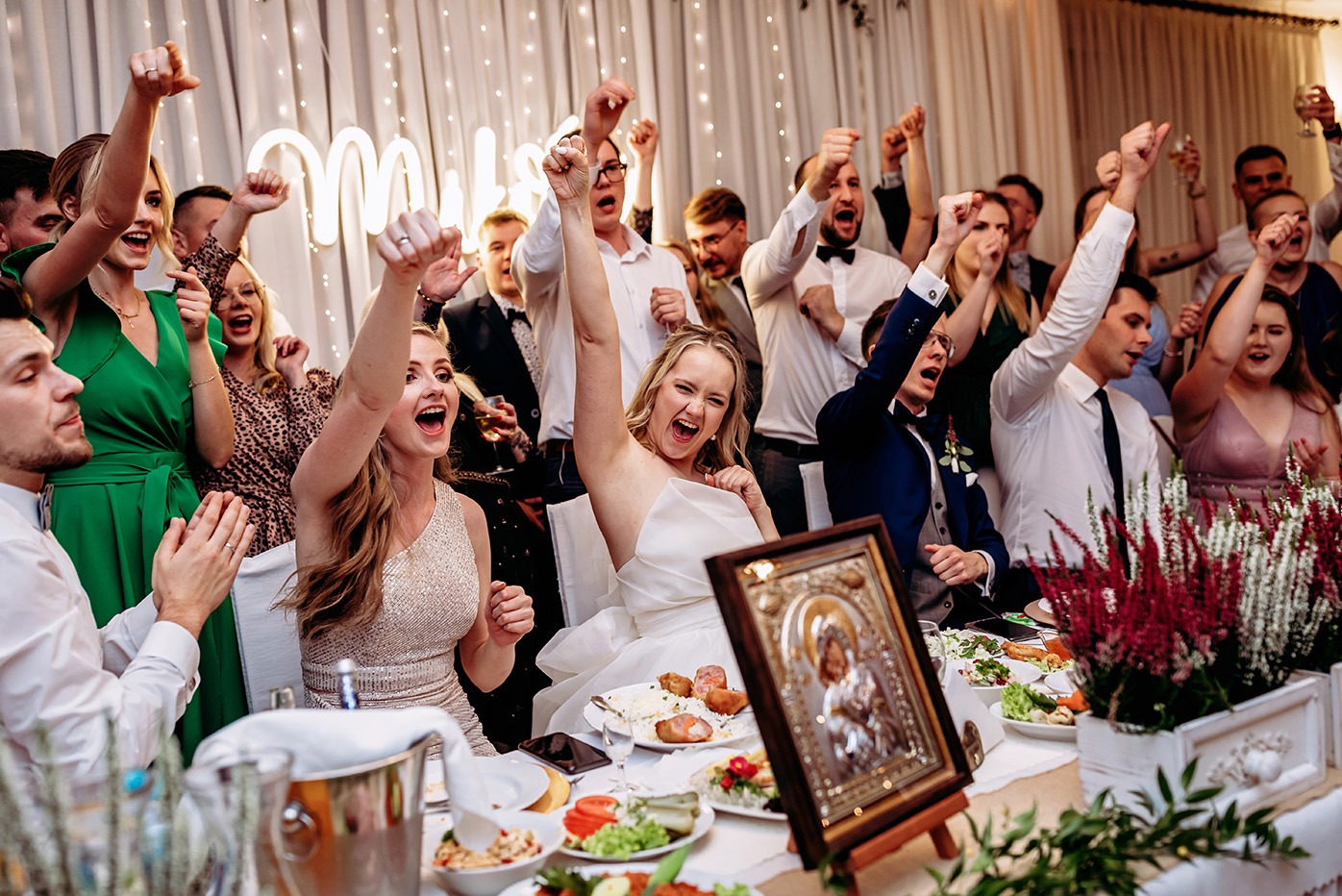 Radosna panna młoda i goście weselni z podniesionymi rękami świętują przy stole z ikoną, na tle dekoracji świetlnych. Ujęcie z bliska, dynamiczne.