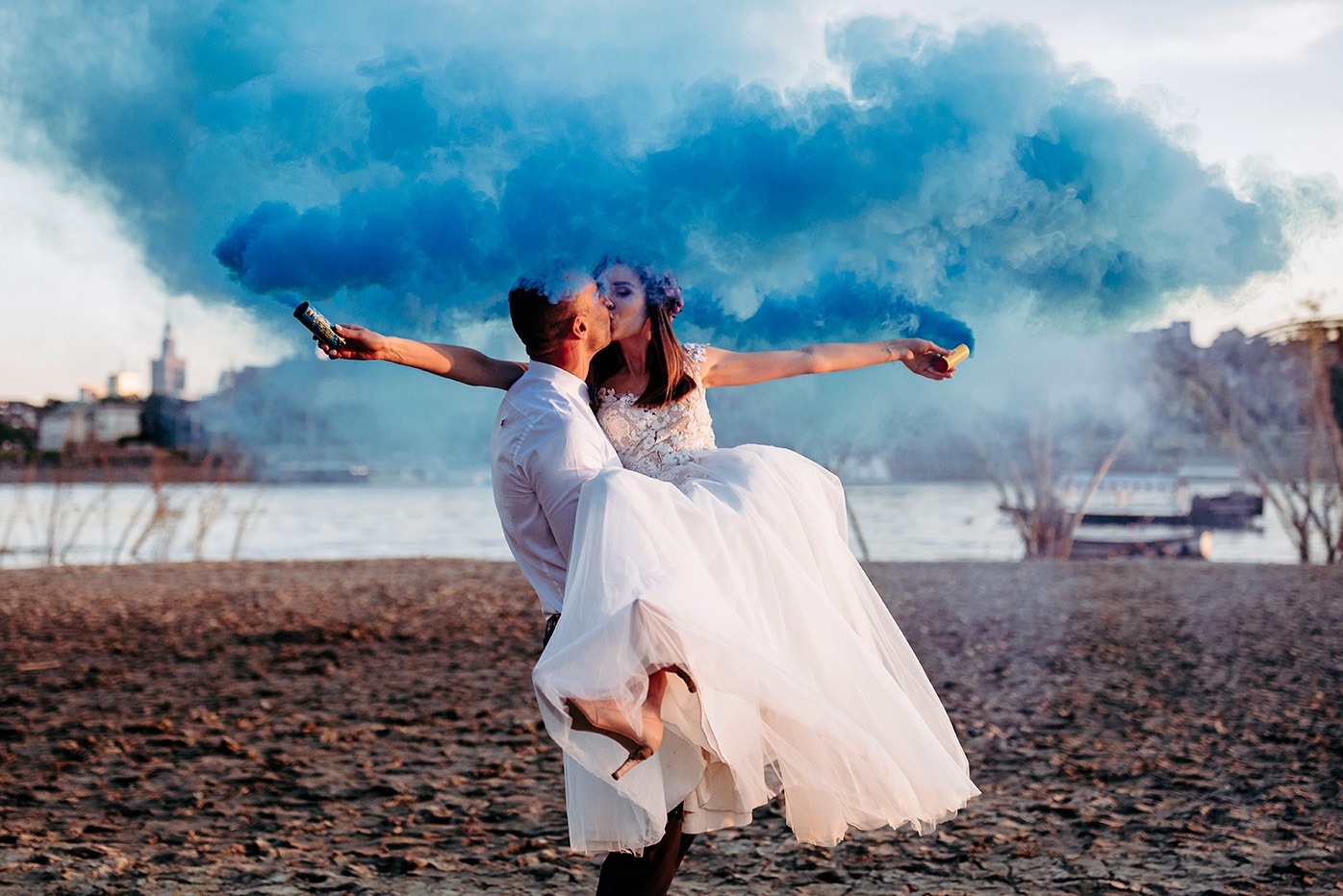 Panna młoda w białej sukni niesiona przez pana młodego na tle rzeki i miasta, z niebieskim dymem z rac.