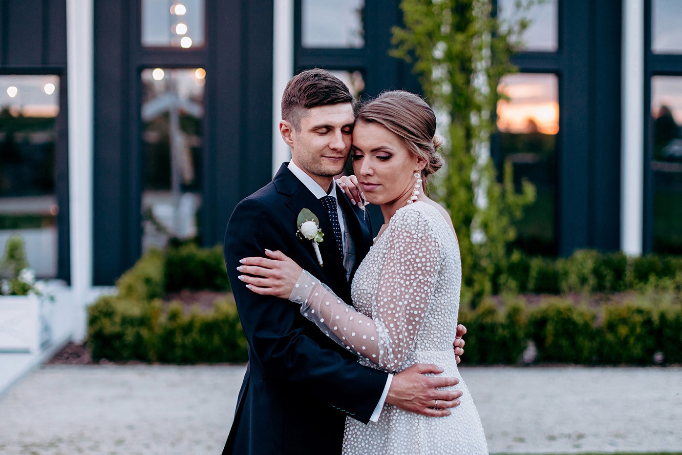 Elegancka para młoda w objęciach na tle nowoczesnej architektury z oknami odbijającymi zachód słońca. Suknia panny młodej z długim rękawem w kropki.