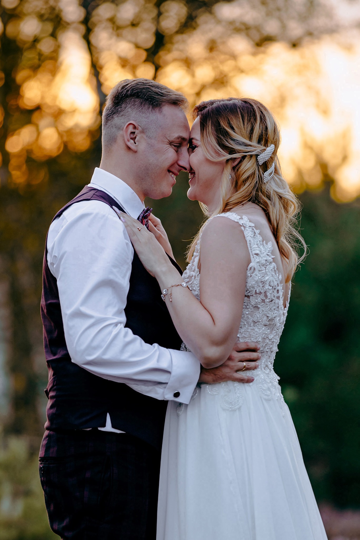 Uśmiechnięta para młoda, zbliżona twarzami w plenerze o zachodzie słońca. Ona w koronkowej sukni, on w bordowej kamizelce i koszuli. Romantyczny moment.