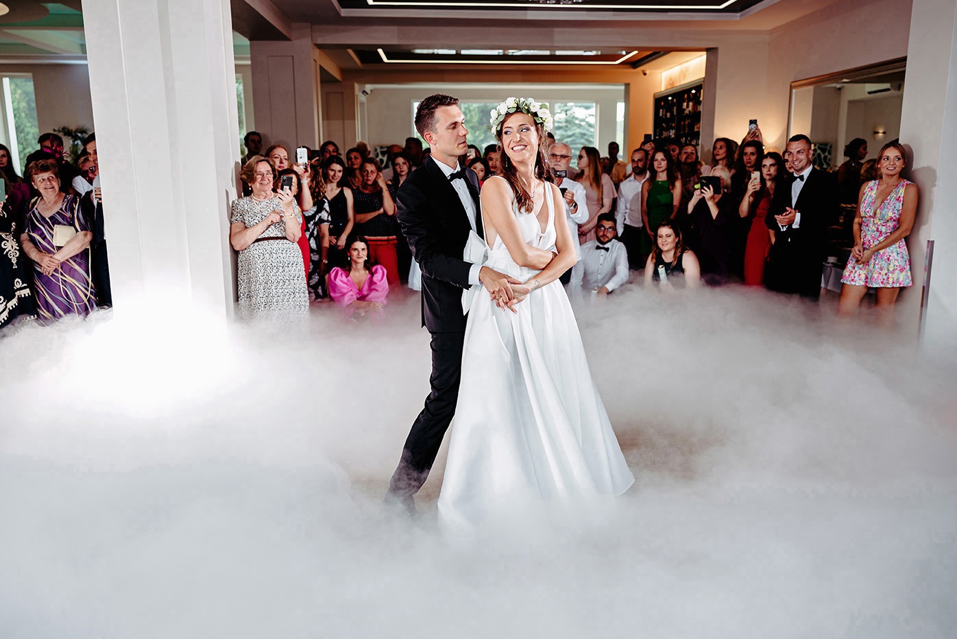 Para młoda w tańcu, otoczona mgłą, na tle gości weselnych. Panna młoda w białej sukni i wianku, pan młody w czarnym smokingu. Uśmiechnięci, w eleganckim wnętrzu.