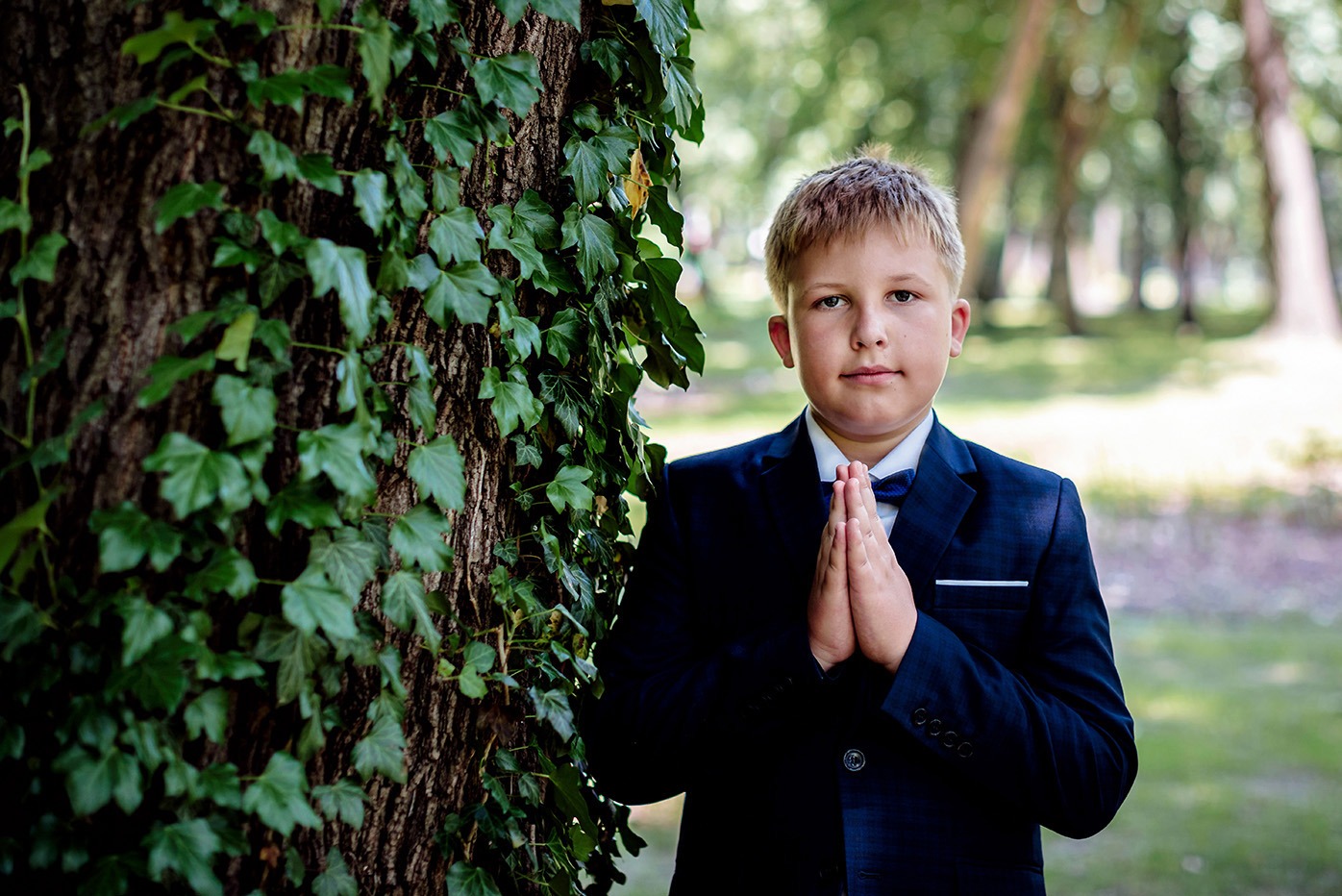 Chłopiec w granatowym garniturze i muszce, z rękami złożonymi do modlitwy, oparty o drzewo obrośnięte bluszczem, w tle rozmyty park.