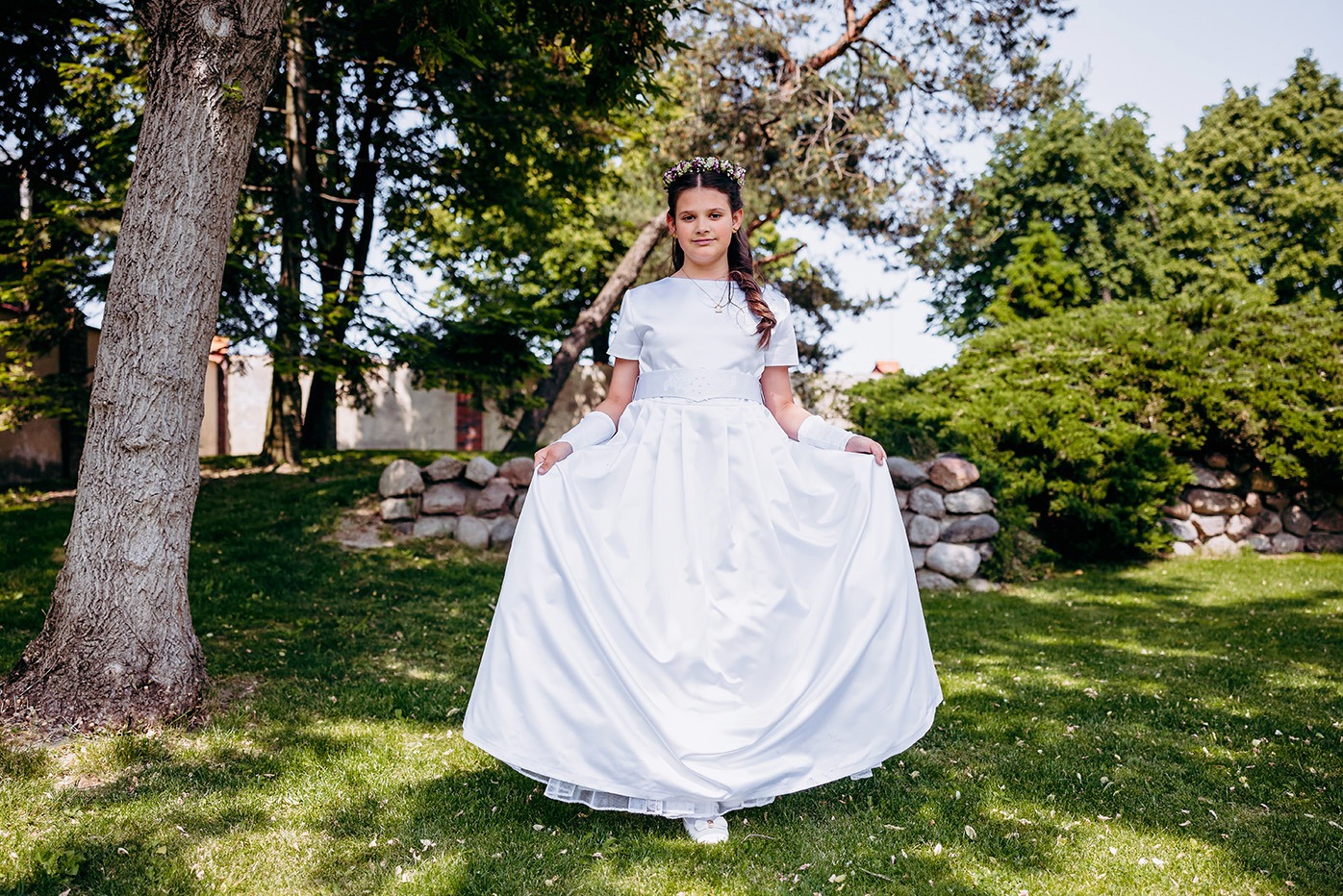 Dziewczynka w białej sukience komunijnej, stojąca na trawie przed kamiennym murkiem, w tle drzewa i budynek. Ujęcie z przodu, w pełnym słońcu.