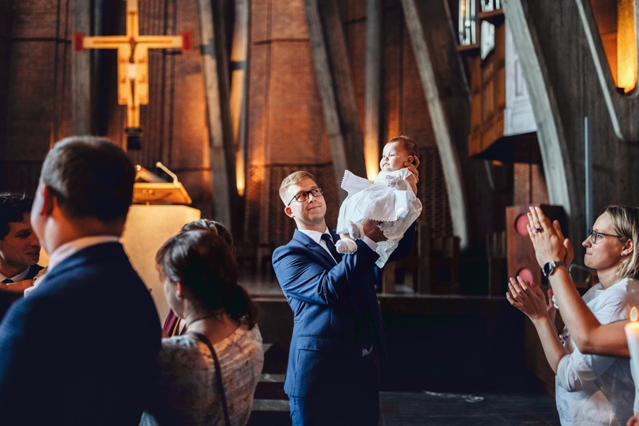 Uśmiechnięty ojciec w garniturze trzyma na rękach ubraną w białą sukienkę córeczkę podczas uroczystości chrztu w kościele. W tle goście i ołtarz.